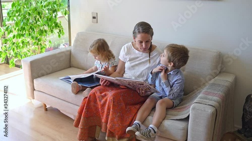 Young mother reading a book to her two children and having fun at home during quarantine