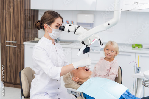 A dentist looking into a woman's mouth through a dental microscope, the woman's daughter is beside her mom waiting for her to complete the check up