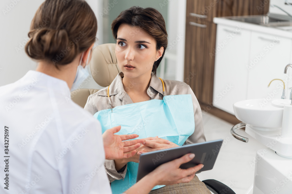 A young worried woman sitting in a dental chair and looking at her doctor, while the dentist is explaining something to her using modern technology