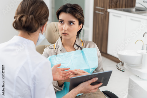 A young worried woman sitting in a dental chair and looking at her doctor  while the dentist is explaining something to her using modern technology
