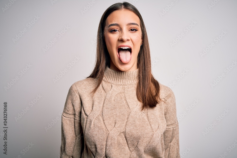 Young beautiful brunette woman wearing casual sweater standing over white background sticking tongue out happy with funny expression. Emotion concept.