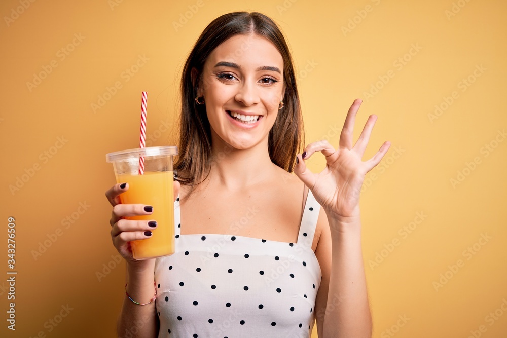 Young beautiful brunette woman drinking healthy orange juice over yellow background doing ok sign with fingers, excellent symbol Young beautiful brunette woman drinking healthy orange juice over yellow background doing ok sign with fingers, excellent symbol