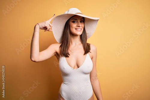 Young beautiful brunette woman on vacation wearing swimsuit and summer hat Smiling pointing to head with one finger, great idea or thought, good memory
