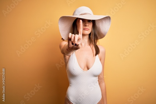 Young beautiful brunette woman on vacation wearing swimsuit and summer hat Pointing with finger up and angry expression, showing no gesture