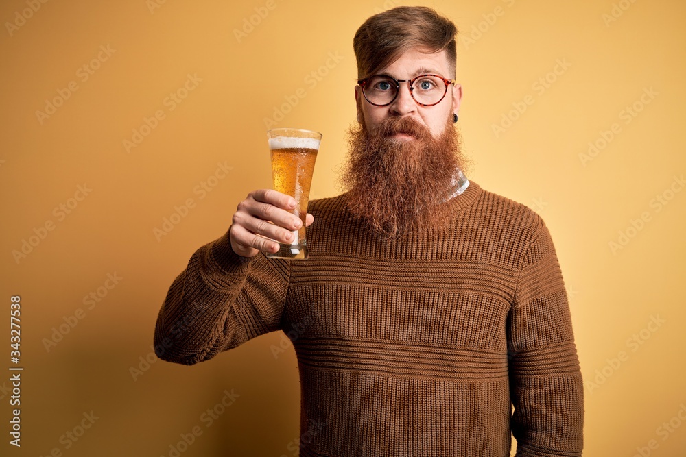 Irish redhead man with beard drinking a glass of refreshing beer over yellow background with a confident expression on smart face thinking serious Irish redhead man with beard drinking a glass of refreshing beer over yellow background with a confident expression on smart face thinking serious