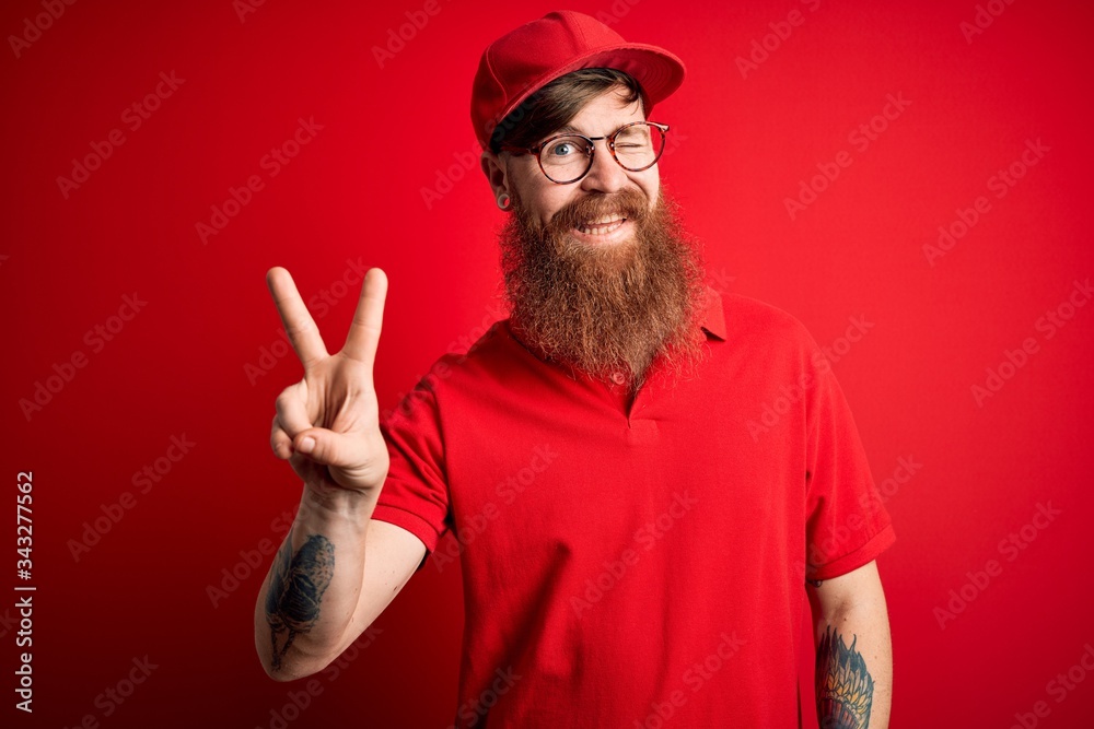 Young handsome delivery man wearing glasses and red cap over isolated background smiling with happy face winking at the camera doing victory sign with fingers. Number two. Young handsome delivery man wearing glasses and red cap over isolated background smiling with happy face winking at the camera doing victory sign with fingers. Number two.