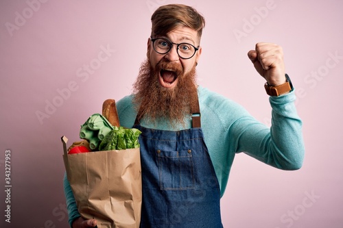 Redhead Irish supermarket worker man with beard wearing apron and holding fresh groceries screaming proud and celebrating victory and success very excited, cheering emotion