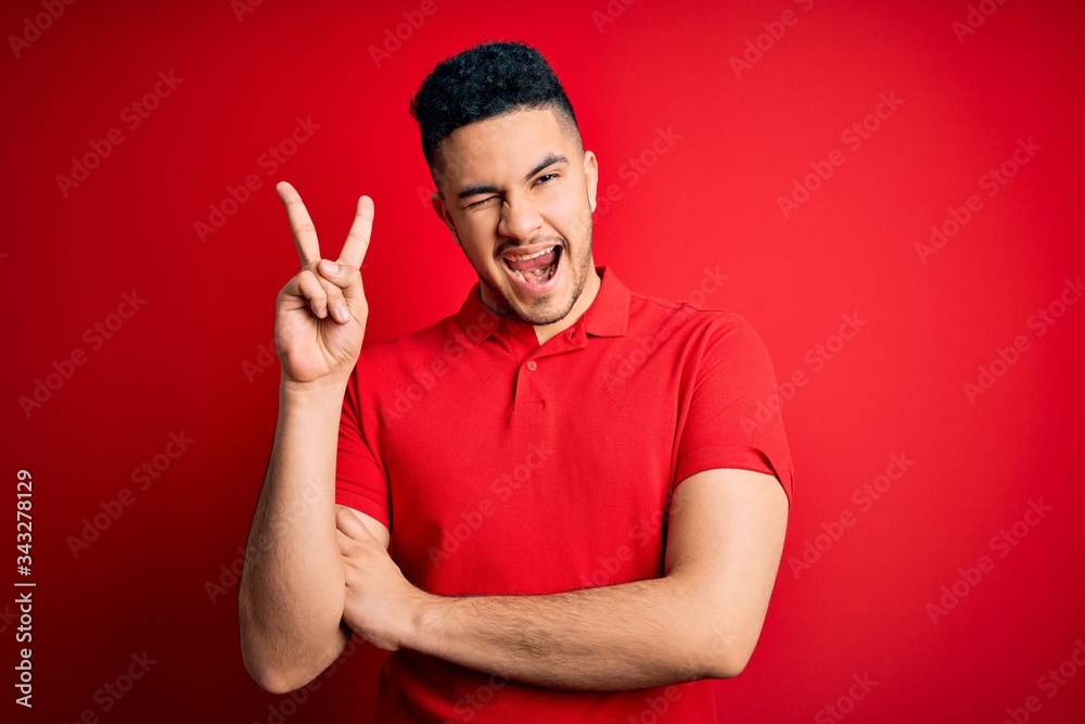 Naklejka premium Young handsome man wearing red casual polo standing over isolated background smiling with happy face winking at the camera doing victory sign. Number two.