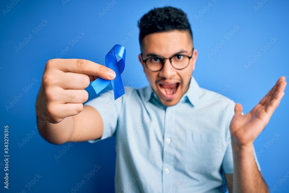 Young handsome man holding blue cancer ribbon standing over isolated background very happy and excited, winner expression celebrating victory screaming with big smile and raised hands Young handsome man holding blue cancer ribbon standing over isolated background very happy and excited, winner expression celebrating victory screaming with big smile and raised hands
