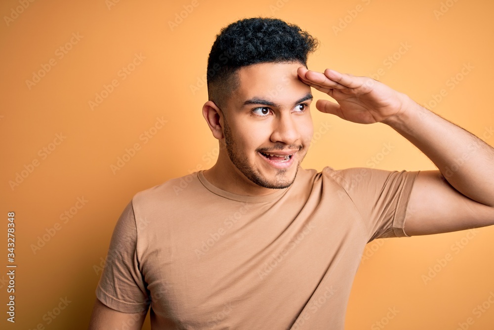 Young handsome man wearing casual t-shirt standing over isolated yellow background very happy and smiling looking far away with hand over head. Searching concept. Young handsome man wearing casual t-shirt standing over isolated yellow background very happy and smiling looking far away with hand over head. Searching concept.