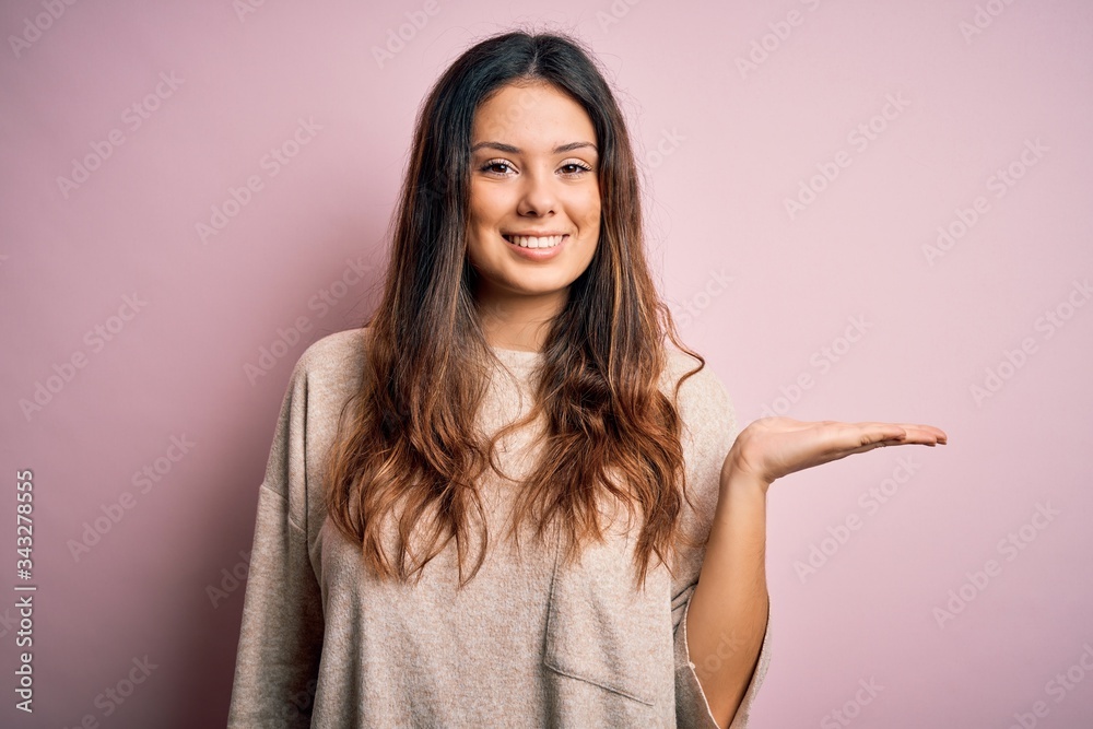Young beautiful brunette woman wearing casual sweater standing over pink background smiling cheerful presenting and pointing with palm of hand looking at the camera.