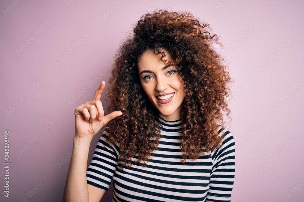 Young beautiful woman with curly hair and piercing wearing casual striped t-shirt smiling and confident gesturing with hand doing small size sign with fingers looking and the camera. Measure concept. Young beautiful woman with curly hair and piercing wearing casual striped t-shirt smiling and confident gesturing with hand doing small size sign with fingers looking and the camera. Measure concept.