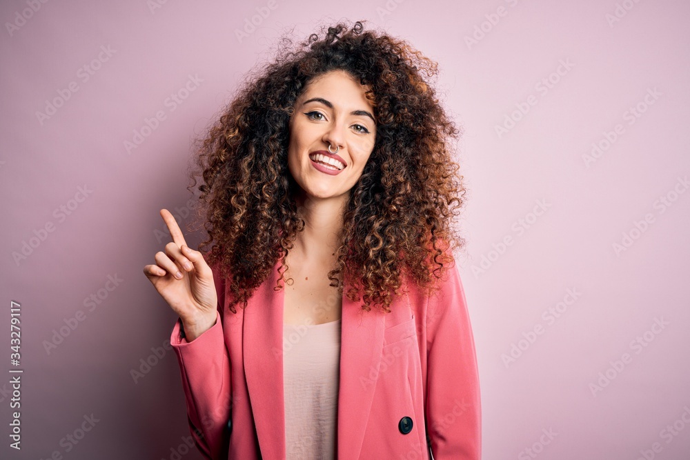 Young beautiful businesswoman with curly hair and piercing wearing elegant jacket with a big smile on face, pointing with hand finger to the side looking at the camera. Young beautiful businesswoman with curly hair and piercing wearing elegant jacket with a big smile on face, pointing with hand finger to the side looking at the camera.