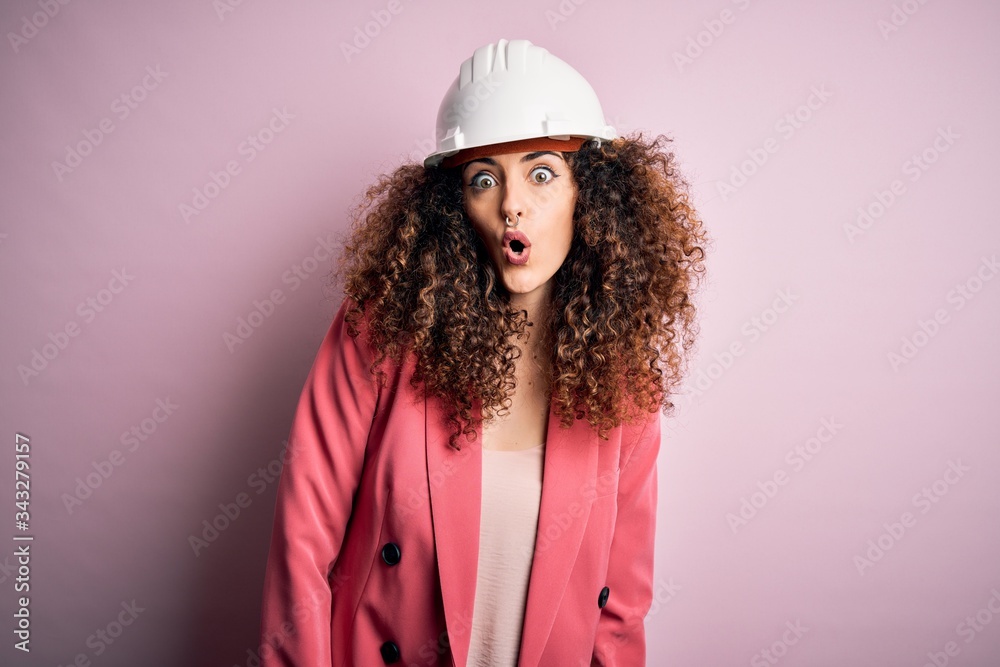 Young beautiful architect woman with curly hair wearing safety helmet over pink background afraid and shocked with surprise expression, fear and excited face.