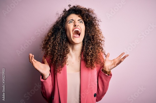 Young beautiful businesswoman with curly hair and piercing wearing elegant jacket crazy and mad shouting and yelling with aggressive expression and arms raised. Frustration concept.