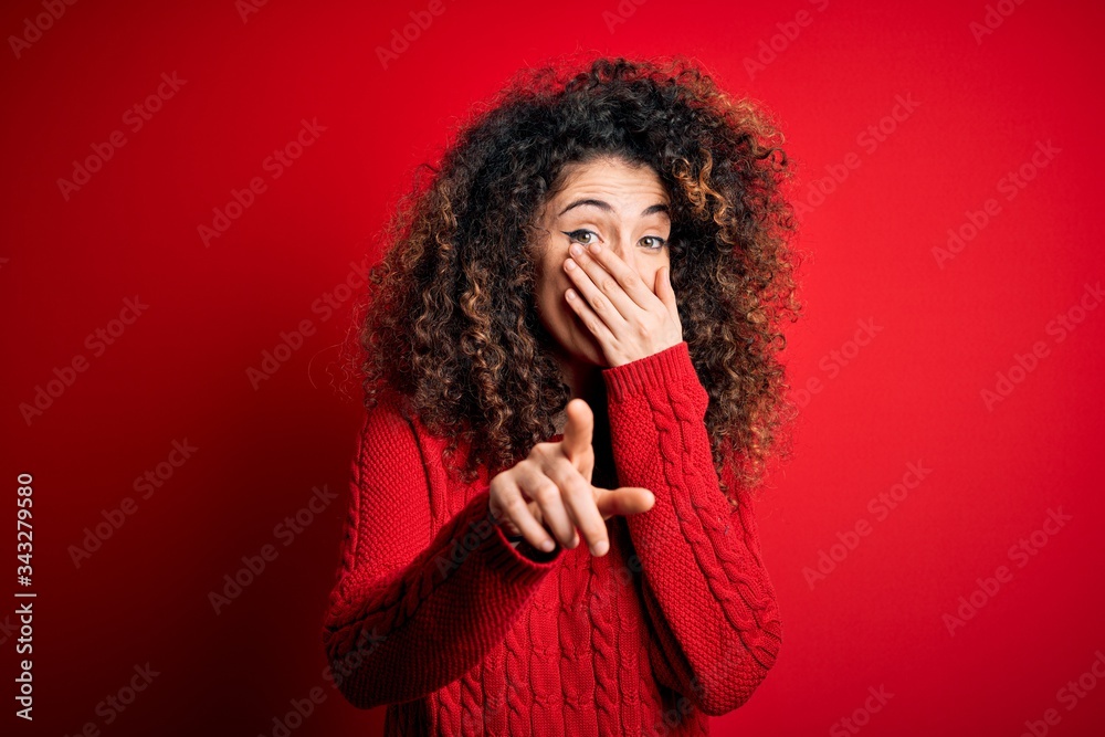 Young beautiful woman with curly hair and piercing wearing casual red sweater laughing at you, pointing finger to the camera with hand over mouth, shame expression Young beautiful woman with curly hair and piercing wearing casual red sweater laughing at you, pointing finger to the camera with hand over mouth, shame expression