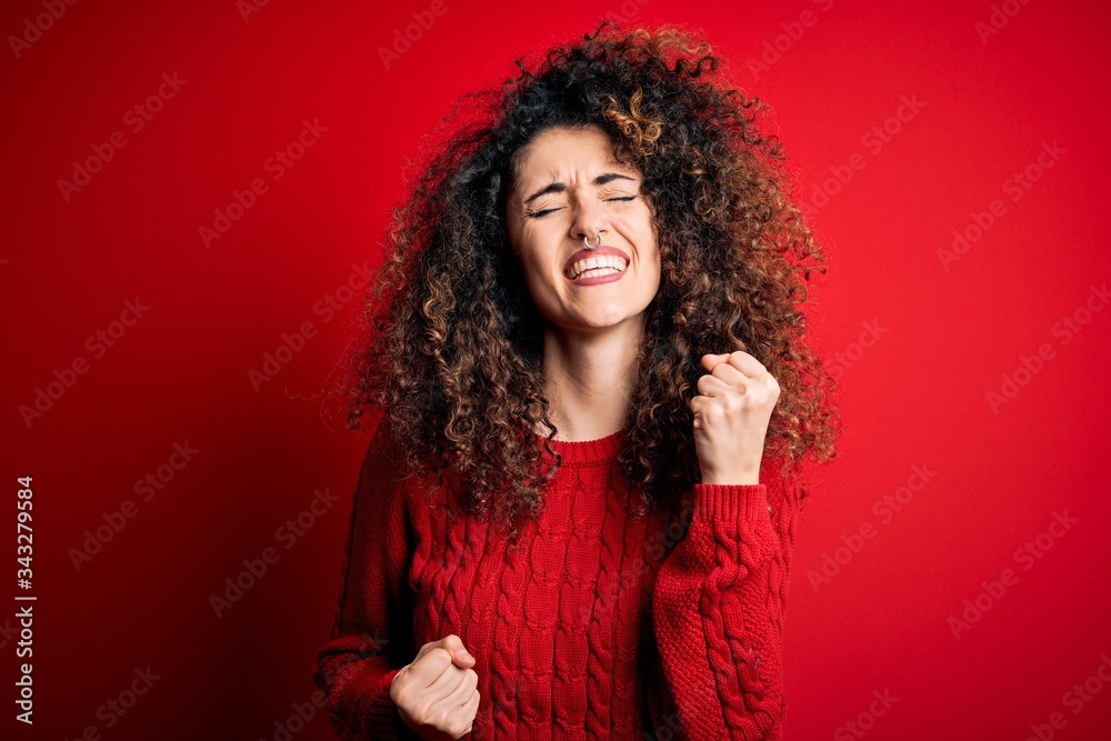 Young beautiful woman with curly hair and piercing wearing casual red sweater celebrating surprised and amazed for success with arms raised and eyes closed. Winner concept. Young beautiful woman with curly hair and piercing wearing casual red sweater celebrating surprised and amazed for success with arms raised and eyes closed. Winner concept.