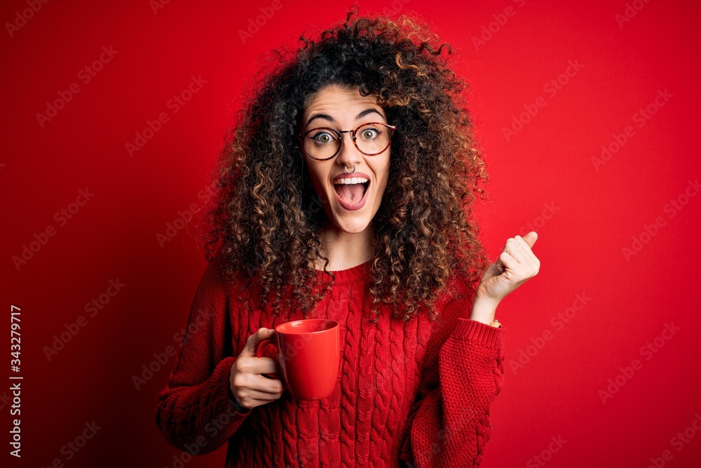 Young beautiful woman with curly hair and piercing drinking red cup of coffee screaming proud and celebrating victory and success very excited, cheering emotion Young beautiful woman with curly hair and piercing drinking red cup of coffee screaming proud and celebrating victory and success very excited, cheering emotion