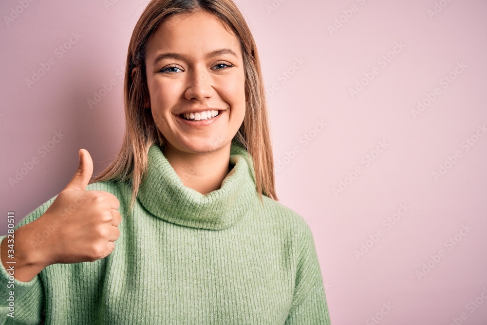 Young beautiful blonde woman wearing winter wool sweater over pink isolated background doing happy thumbs up gesture with hand. Approving expression looking at the camera showing success. Young beautiful blonde woman wearing winter wool sweater over pink isolated background doing happy thumbs up gesture with hand. Approving expression looking at the camera showing success.