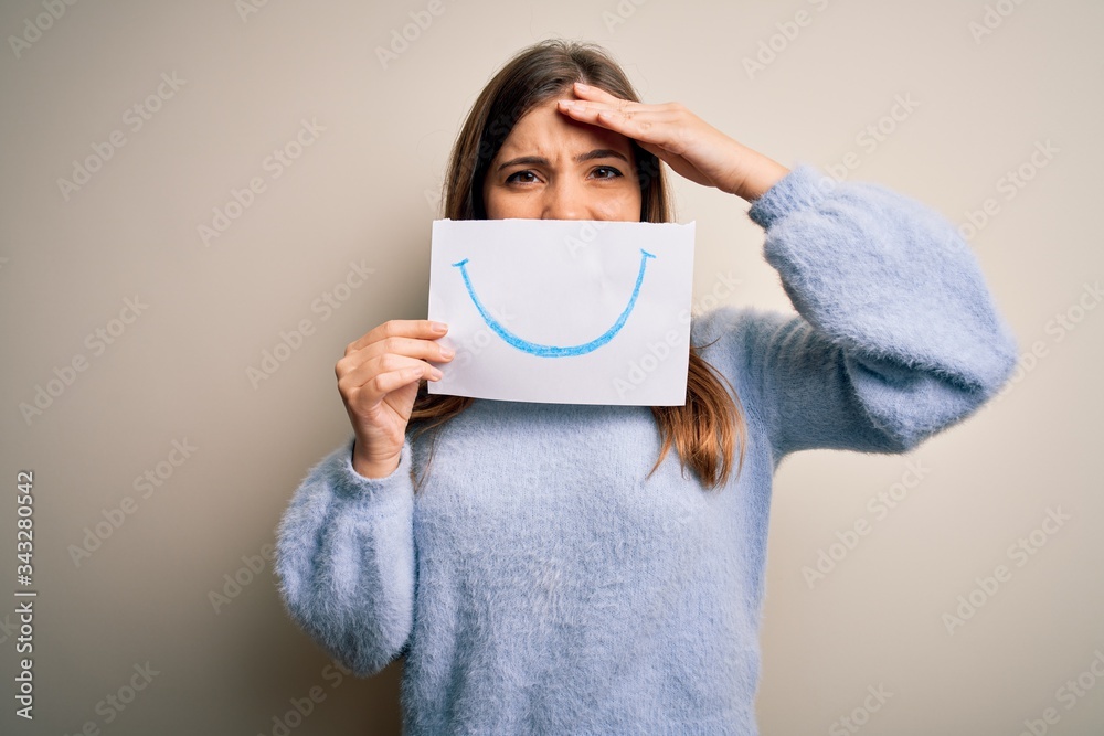 Young blonde woman holding funny smile drawing on mouth as happy expression stressed with hand on head, shocked with shame and surprise face, angry and frustrated. Fear and upset for mistake. Young blonde woman holding funny smile drawing on mouth as happy expression stressed with hand on head, shocked with shame and surprise face, angry and frustrated. Fear and upset for mistake.