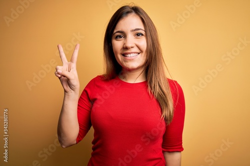 Beautiful young woman wearing casual red t-shirt over yellow isolated background smiling with happy face winking at the camera doing victory sign. Number two.
