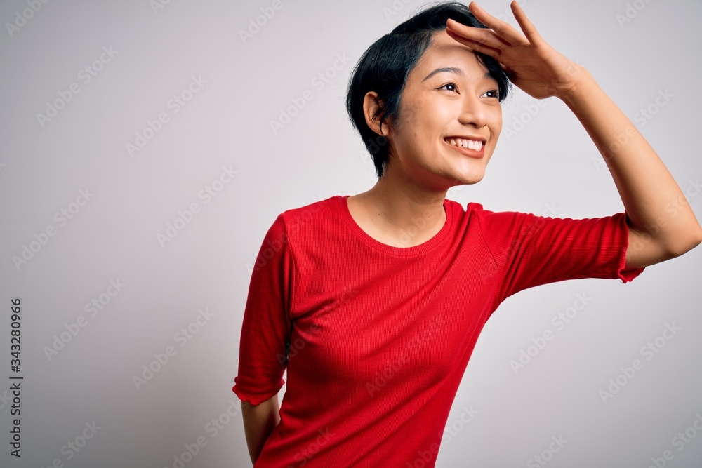 Young beautiful asian girl wearing casual red t-shirt standing over isolated white background very happy and smiling looking far away with hand over head. Searching concept. Young beautiful asian girl wearing casual red t-shirt standing over isolated white background very happy and smiling looking far away with hand over head. Searching concept.