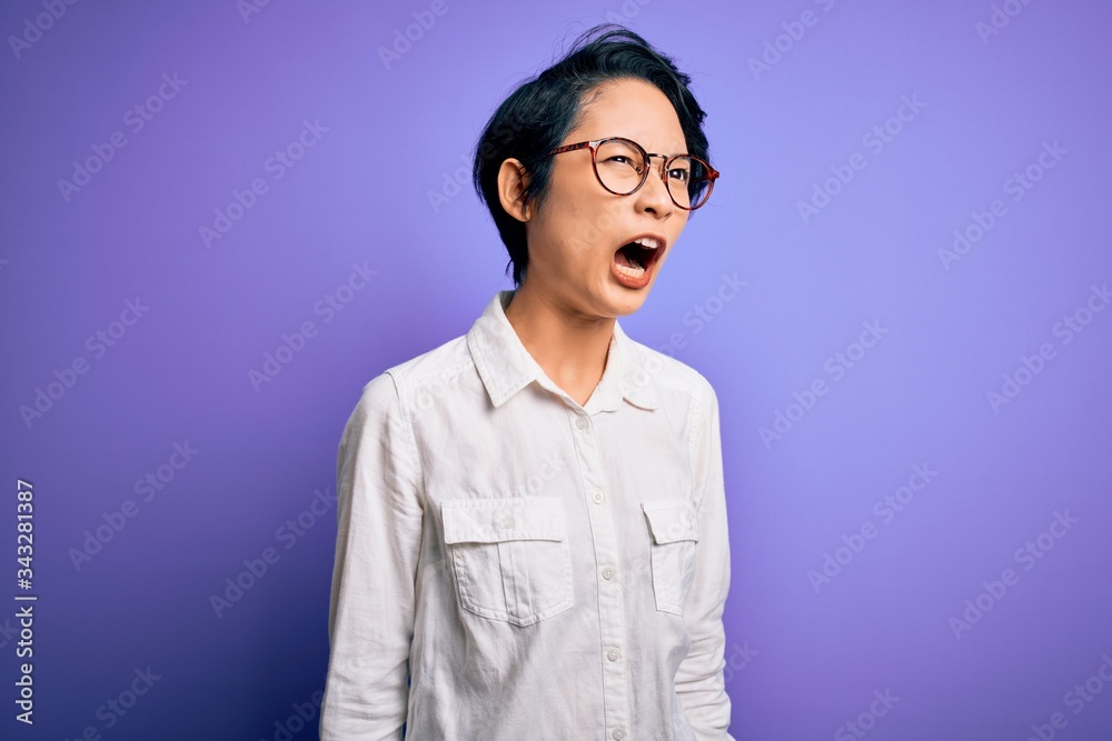 Young beautiful asian girl wearing casual shirt and glasses standing over purple background angry and mad screaming frustrated and furious, shouting with anger. Rage and aggressive concept.