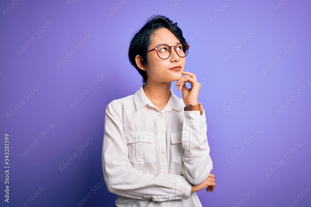 Young beautiful asian girl wearing casual shirt and glasses standing over purple background with hand on chin thinking about question, pensive expression. Smiling with thoughtful face. Doubt concept. Young beautiful asian girl wearing casual shirt and glasses standing over purple background with hand on chin thinking about question, pensive expression. Smiling with thoughtful face. Doubt concept.