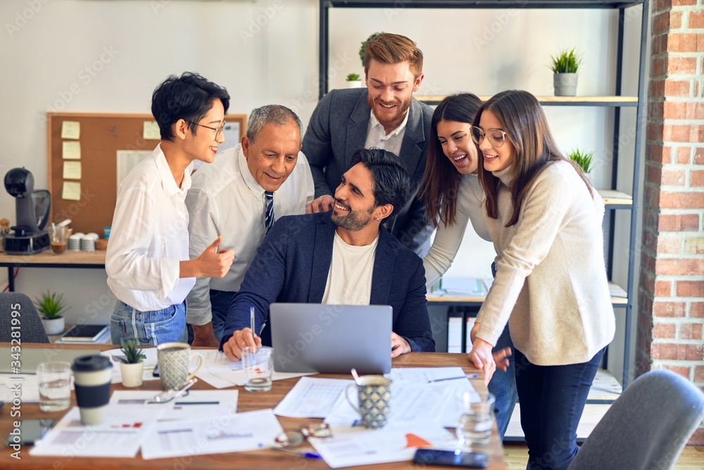 Group of business workers smiling happy and confident. One of them sitting and partners standing around. Working together with smile on face looking at the laptop at the office Group of business workers smiling happy and confident. One of them sitting and partners standing around. Working together with smile on face looking at the laptop at the office