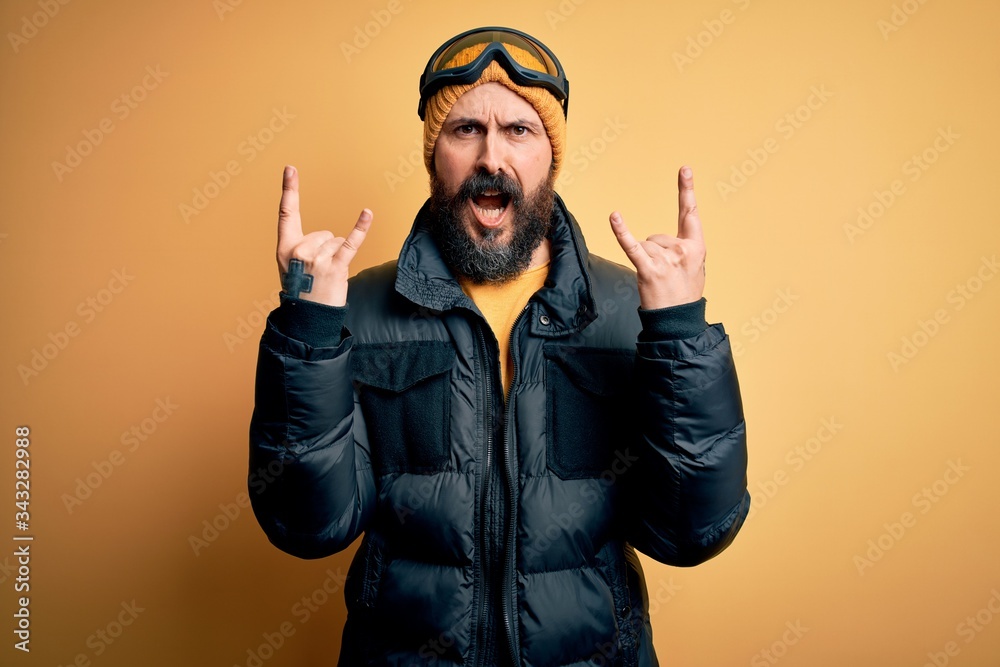 Handsome skier bald man with beard skiing wearing snow sportswear and ski goggles shouting with crazy expression doing rock symbol with hands up. Music star. Heavy concept. Handsome skier bald man with beard skiing wearing snow sportswear and ski goggles shouting with crazy expression doing rock symbol with hands up. Music star. Heavy concept.