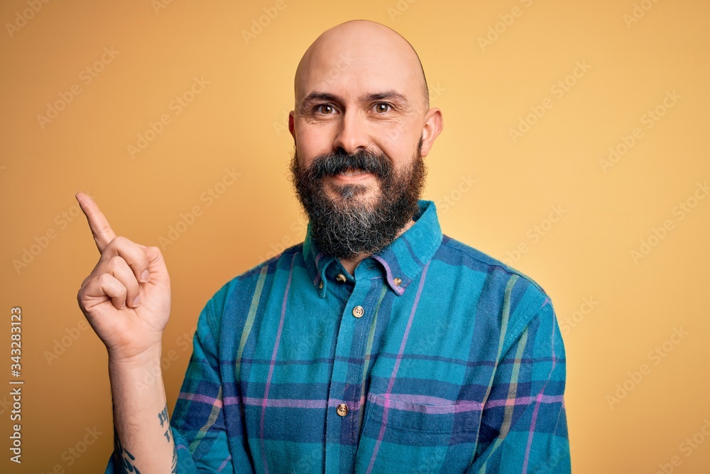 Handsome bald man with beard wearing casual shirt standing over isolated yellow background with a big smile on face, pointing with hand and finger to the side looking at the camera. Handsome bald man with beard wearing casual shirt standing over isolated yellow background with a big smile on face, pointing with hand and finger to the side looking at the camera.