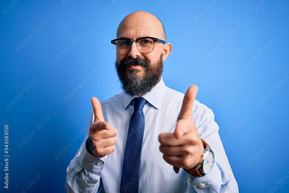 Handsome business bald man with beard wearing elegant tie and glasses over blue background pointing fingers to camera with happy and funny face. Good energy and vibes. Handsome business bald man with beard wearing elegant tie and glasses over blue background pointing fingers to camera with happy and funny face. Good energy and vibes.