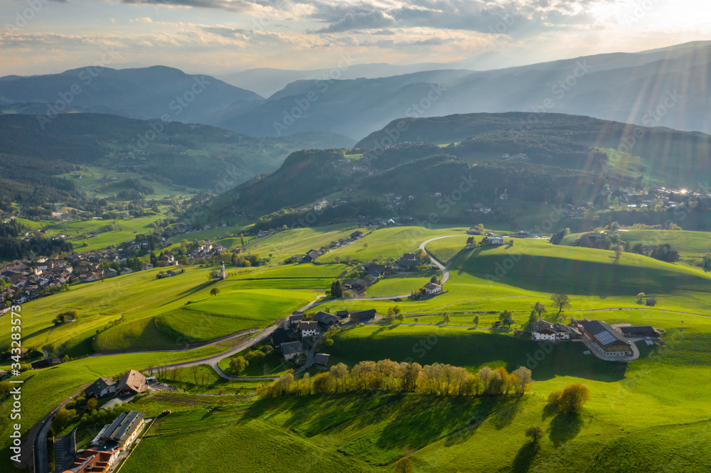 Aerial view of improbable green meadows of Italian Alps, green slopes of the mountains, Bolzano, huge clouds over a valley, roof tops of houses, Dolomites on background, sunshines through clouds