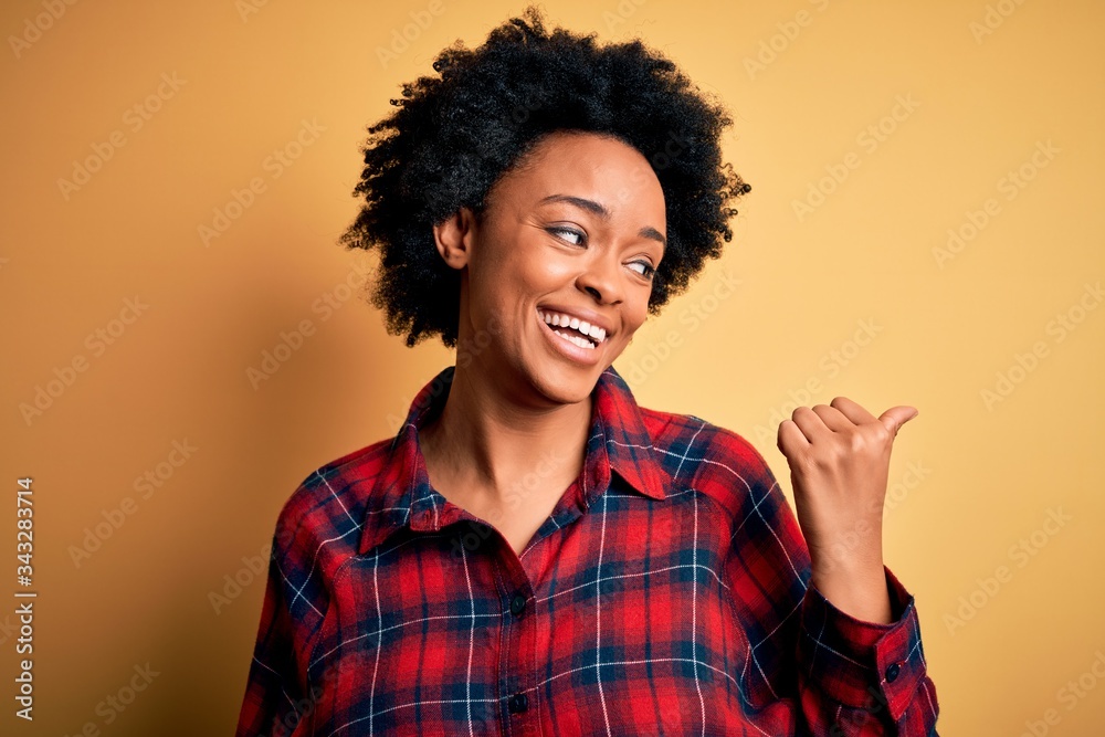 Young beautiful African American afro woman with curly hair wearing casual shirt smiling with happy face looking and pointing to the side with thumb up.