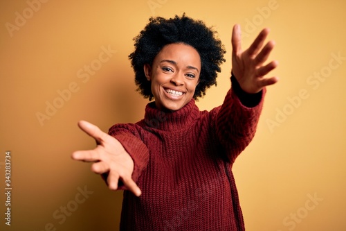 Young beautiful African American afro woman with curly hair wearing casual turtleneck sweater looking at the camera smiling with open arms for hug. Cheerful expression embracing happiness.