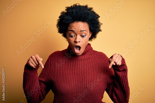 Young beautiful African American afro woman with curly hair wearing casual turtleneck sweater Pointing down with fingers showing advertisement, surprised face and open mouth © Krakenimages.com
