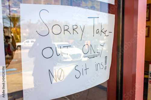 Handwritten sign at restaurant stating Sorry Take-Out Only No Sit Ins due to coronavirus pandemic. Person and parking lot are reflected in the window