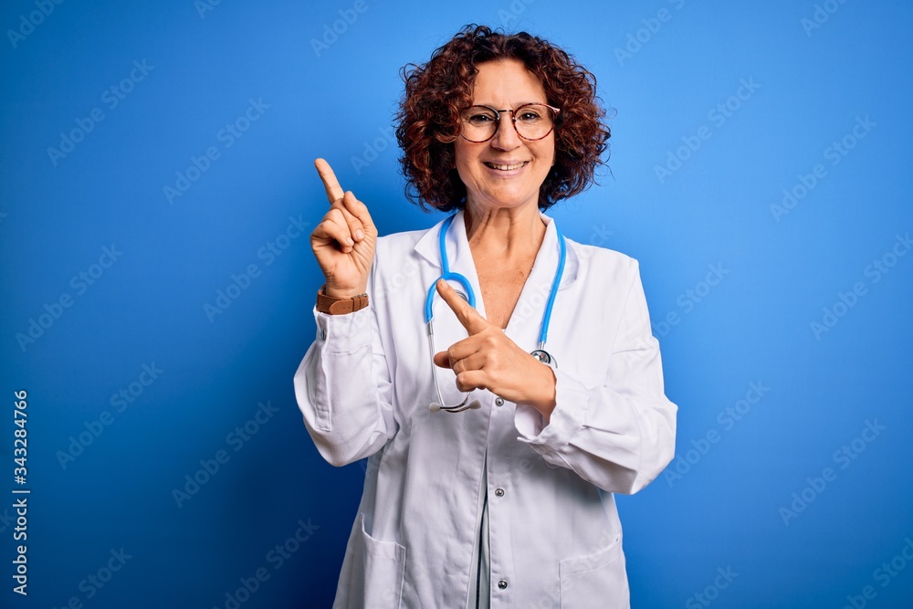 Middle age curly hair doctor woman wearing coat and stethoscope over blue background smiling and looking at the camera pointing with two hands and fingers to the side. Middle age curly hair doctor woman wearing coat and stethoscope over blue background smiling and looking at the camera pointing with two hands and fingers to the side.