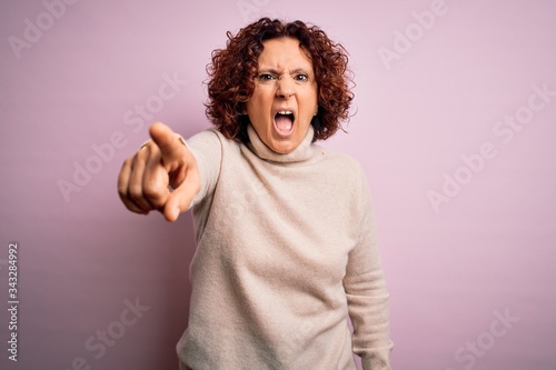 Middle age beautiful curly hair woman wearing casual turtleneck sweater over pink background pointing displeased and frustrated to the camera, angry and furious with you