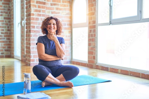 Middle age beautiful sportswoman wearing sportswear sitting on mat practicing yoga at home looking confident at the camera smiling with crossed arms and hand raised on chin. Thinking positive.