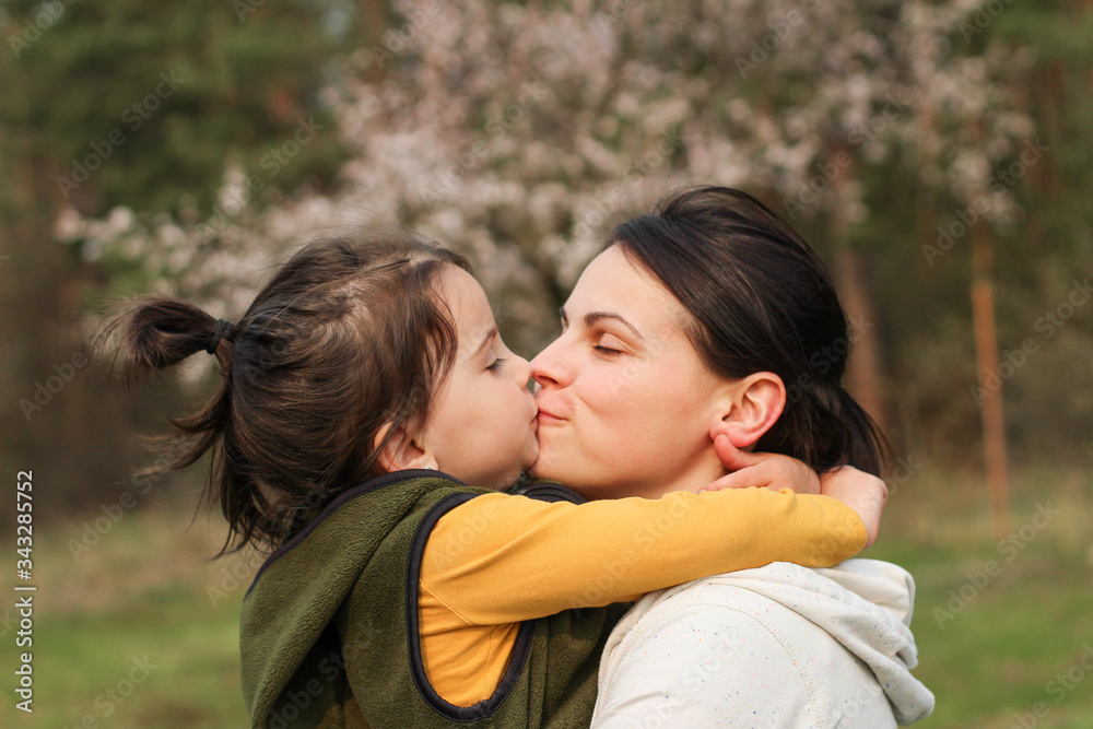 Beautiful mother and daughter hugging and kissing in casual clothes. Warm baby hug on the background of a blurry flowering tree Beautiful mother and daughter hugging and kissing in casual clothes. Warm baby hug on the background of a blurry flowering tree