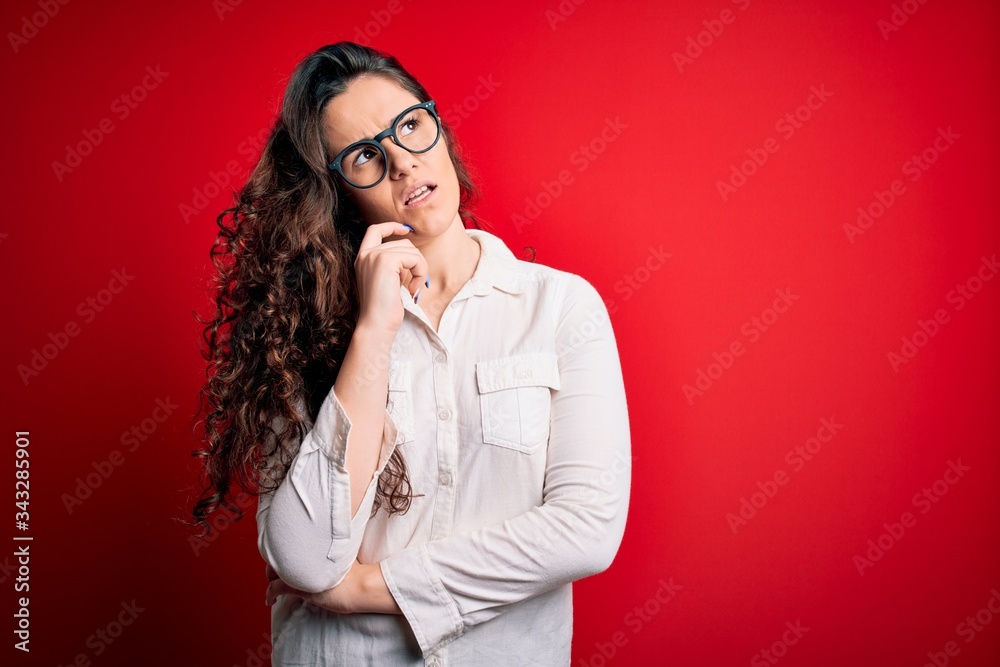 Young beautiful woman with curly hair wearing shirt and glasses over red background with hand on chin thinking about question, pensive expression. Smiling with thoughtful face. Doubt concept. Young beautiful woman with curly hair wearing shirt and glasses over red background with hand on chin thinking about question, pensive expression. Smiling with thoughtful face. Doubt concept.
