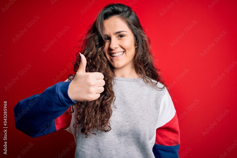 Young beautiful woman with curly hair wearing casual sweatshirt over isolated red background doing happy thumbs up gesture with hand. Approving expression looking at the camera showing success. Young beautiful woman with curly hair wearing casual sweatshirt over isolated red background doing happy thumbs up gesture with hand. Approving expression looking at the camera showing success.