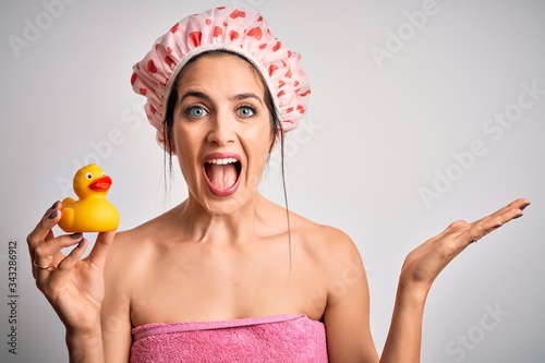 Young brunette woman with blue eyes wearing bath towel and shower cap holding duck toy very happy and excited, winner expression celebrating victory screaming with big smile and raised hands