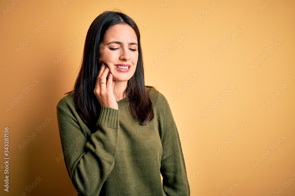 Young brunette woman with blue eyes wearing green casual sweater over yellow background touching mouth with hand with painful expression because of toothache or dental illness on teeth. Dentist Young brunette woman with blue eyes wearing green casual sweater over yellow background touching mouth with hand with painful expression because of toothache or dental illness on teeth. Dentist
