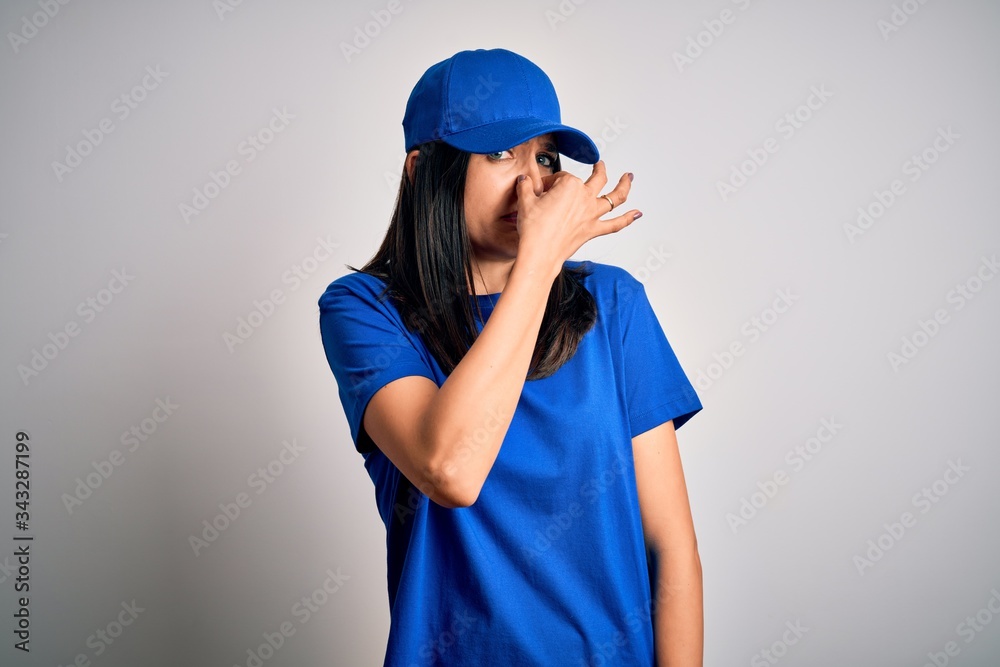 Young delivery woman with blue eyes wearing cap standing over blue background smelling something stinky and disgusting, intolerable smell, holding breath with fingers on nose. Bad smell Young delivery woman with blue eyes wearing cap standing over blue background smelling something stinky and disgusting, intolerable smell, holding breath with fingers on nose. Bad smell