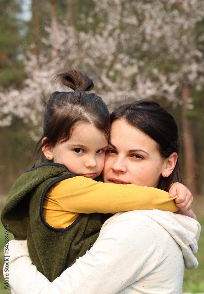 Beautiful mother and daughter hugging and kissing in casual clothes. Warm baby hug on the background of a blurry flowering tree Beautiful mother and daughter hugging and kissing in casual clothes. Warm baby hug on the background of a blurry flowering tree