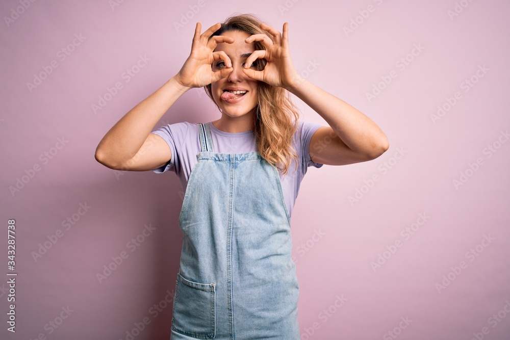 Young beautiful blonde woman wearing casual denim overalls standing over pink background doing ok gesture like binoculars sticking tongue out, eyes looking through fingers. Crazy expression. Young beautiful blonde woman wearing casual denim overalls standing over pink background doing ok gesture like binoculars sticking tongue out, eyes looking through fingers. Crazy expression.