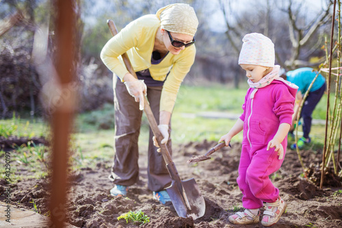 Young woman working in garden while her little daughter playing with spade beside her