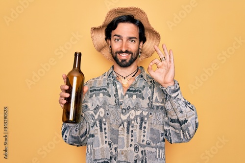 Young handsome hispanic bohemian hipster man with bear drinking from a empty wine bottle doing ok sign with fingers, excellent symbol